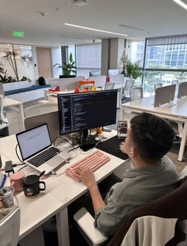 A young man working at a standing desk in a bright modern office, using a laptop with code displayed on the screen, surrounded by plants and colleagues.
