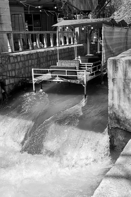 A black and white photograph of turbulent floodwaters rushing past metal bed frames sitting against a concrete wall outside a building.