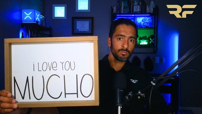 A bearded man in a black shirt sits in a blue-lit room holding a whiteboard that reads "I LOVE YOU MUCCHO" in handwritten black text. Behind him, gaming-themed lights and a framed certificate are visible, with the "RF" logo displayed in the upper right corner.