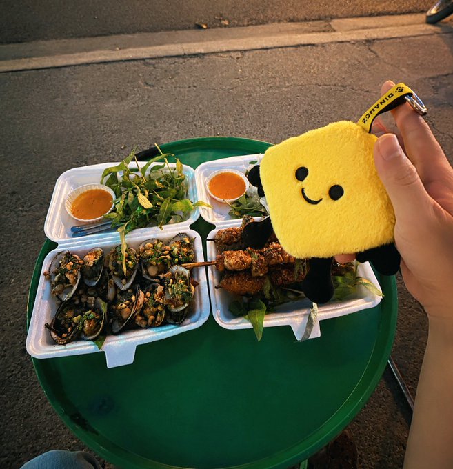 A hand holds a yellow smiley-face plushie above a green table laden with styrofoam containers of seafood — including mussels and meat skewers — served with dipping sauce and fresh herbs.