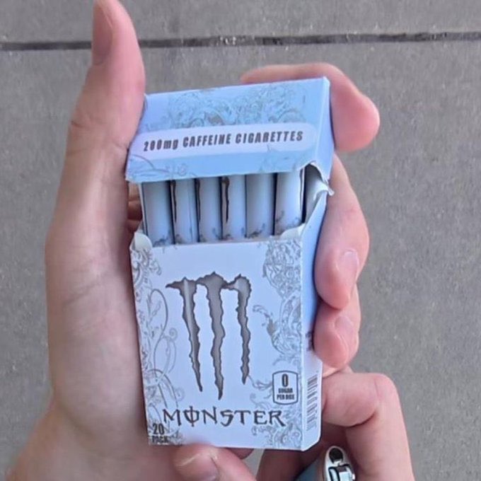 A hand holds an open pack of 'MONSTER' brand '200mg CAFFEINE CIGARETTES,' revealing white filtered cigarettes inside a light blue decorative box.