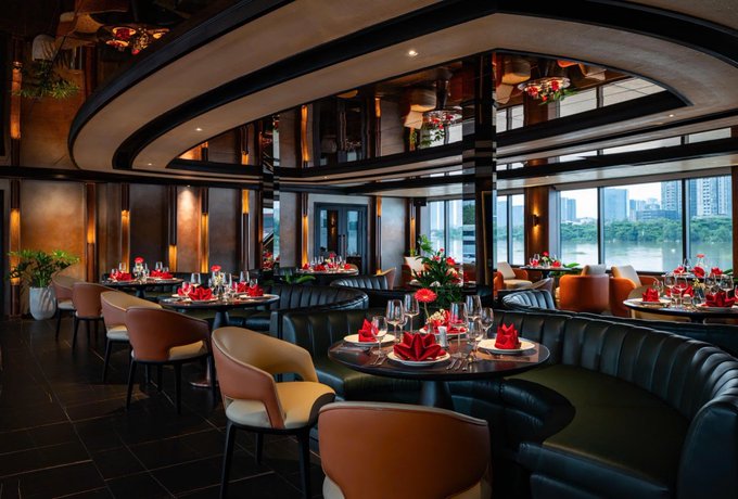 An elegant, upscale restaurant interior with dark leather booths, round tables set with red napkins, and large windows offering a waterfront city view.