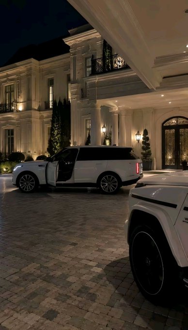 A nighttime photograph showing a luxury white SUV parked at the entrance of an illuminated mansion with classical architecture and a portico.