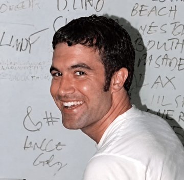 A smiling man in a white t-shirt standing in front of a white wall covered in handwritten text and graffiti.