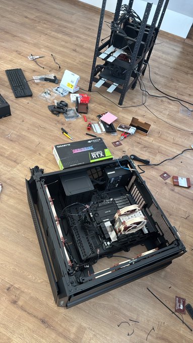 A partially assembled PC build sits on a wooden floor surrounded by scattered components, tools, and a Gigabyte GeForce RTX graphics card box, with a rack of tangled cables towering in the background—capturing the typical mid-build chaos of a tight deadline challenge.