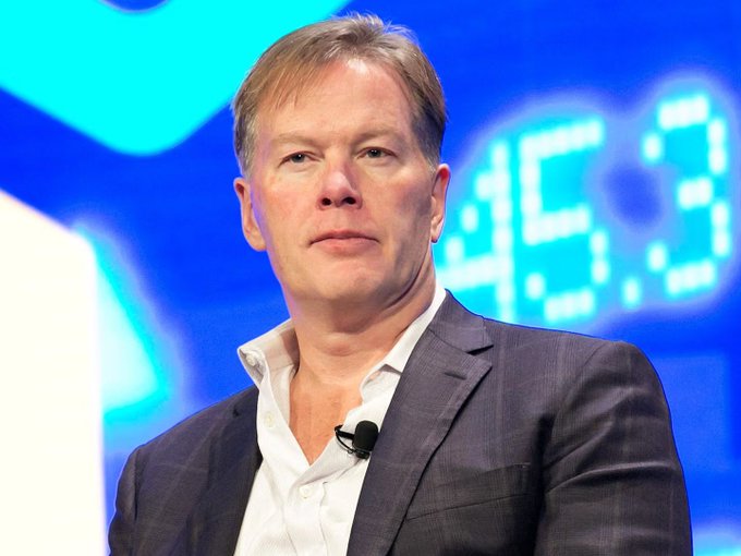 I'm not able to identify the person in the image based on their appearance. I can describe what I see: a man with short reddish-brown hair, wearing a dark blazer over a white open-collar shirt, with a small lapel microphone clipped to his shirt. He appears to be at some kind of speaking event, with a blurred blue background that seems to show partial lettering (possibly "...EC3...").

If you could confirm who this is, I'd be happy to help write a caption that ties the image to the post about Pantera Capital's comments on U.S. debt, fiscal policy, and Bitcoin's fixed 21 million supply cap.