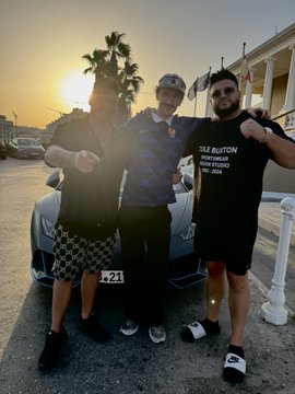 Three young men pose together at sunset near a parked sports car, with one wearing a black "ISLE BURTON" t-shirt and another wearing checkered shorts, appearing to be tourists enjoying a sunny day by the waterfront, likely in Dragonara (Malta). The casual celebratory vibe matches the post's excitement about a good day and plans to "bless" followers by sending crypto to shared SOL addresses.