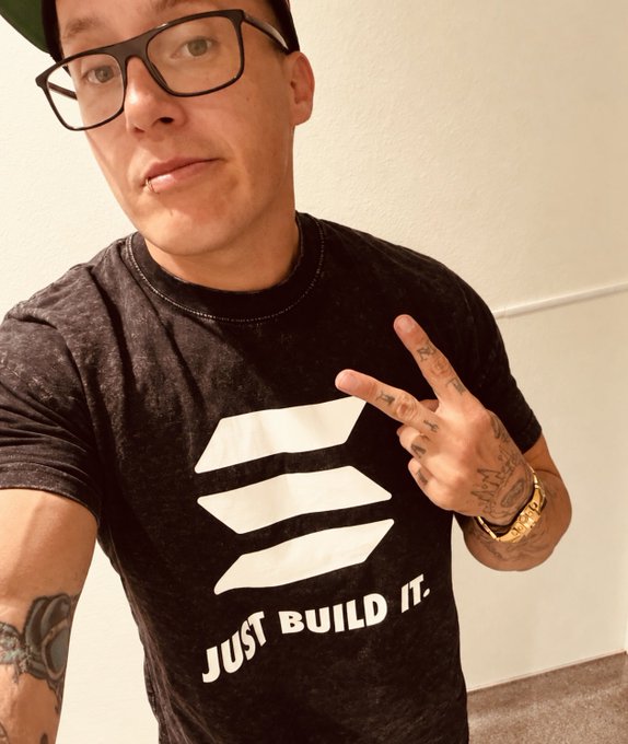 A selfie shows a person with glasses and a lip piercing wearing a black t-shirt featuring white stripes and the text 'JUST BUILD IT.', flashing a peace sign with tattooed hands.