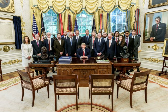 President Donald Trump sits at the Resolute Desk in the Oval Office surrounded by members of his cabinet and senior White House staff, with a portrait of Ronald Reagan visible on the wall.