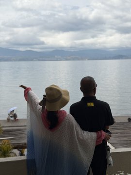 The image shows a man and woman standing together, with the man wearing sunglasses and holding his arm out towards the water. They are both looking at something in the distance, possibly enjoying their time by the lake or watching boats passing by. The scene is set against an overcast sky, which adds to the atmosphere of relaxation and leisurely activities.