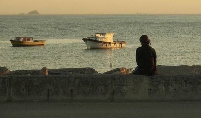 A serene coastal scene at sunset with a person sitting on a stone wall watching two boats anchored in calm waters, with a small island visible on the horizon.