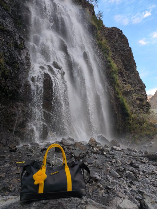 A black and yellow Lancel duffle bag sits on rocky terrain in front of a powerful waterfall cascading down a steep cliff face.