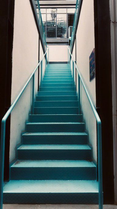A narrow industrial staircase with teal-painted steps and railings leading upward to a doorway, framed by white walls and dark vertical columns in a factory-like setting.