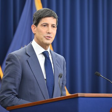 A man in a blue suit and tie stands at a podium speaking, with a blue flag visible behind him in what appears to be an official press briefing.