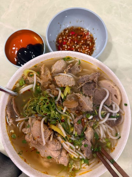 A bowl of Vietnamese noodle soup (likely bún bò) topped with sliced beef, pork, bean sprouts, and green onions, served with two small side dishes of dipping sauces.