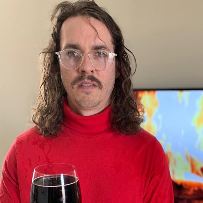 A man with long curly hair, glasses, and a mustache wears a red turtleneck and holds a glass of red wine.