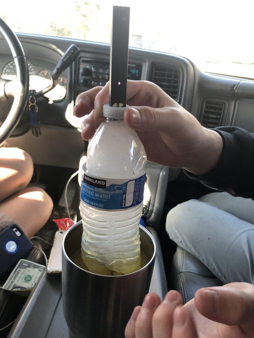 A young man holds a Kirkland purified water bottle with a refractometer inserted into the cap, testing whether the liquid inside is actually water—all while sitting in the driver's seat of a car. The image humorously contrasts the meme's joke about 18-year-olds making millions in crypto, suggesting that at 18, this person was instead preoccupied with mundane (and possibly paranoid) tasks like verifying if his bottled water had been tampered with.