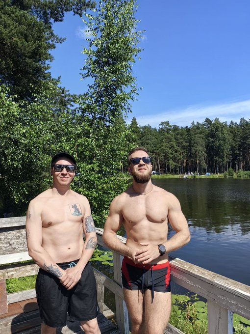 Two shirtless men posing together on a wooden deck by a lake surrounded by trees, with one wearing black swim trunks and the other in red and black trunks with a watch.