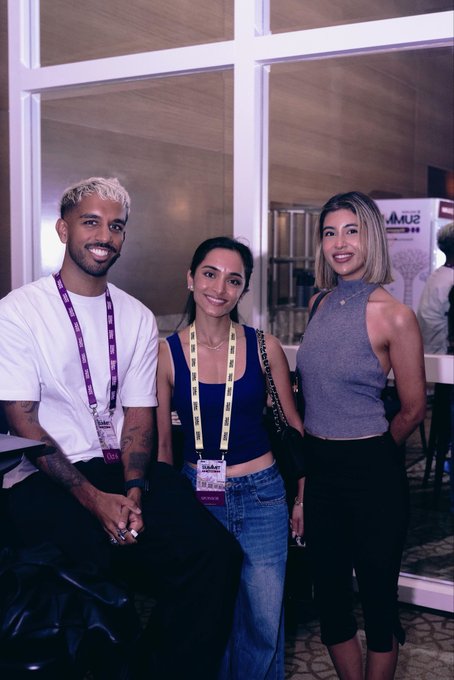 Three attendees pose together at what appears to be a Solana-related event, with a man in a white t-shirt, a woman in a dark tank top, and a woman in a gray halter top, all wearing event lanyards in front of a "SUMMIT" sign — fitting the "Solana cinematic universe" theme of gathering crypto enthusiasts and builders at a conference.