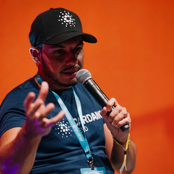 A man wearing a black cap and a blue t-shirt with a dot pattern speaks into a microphone at a BlockDAG event, wearing a teal lanyard against an orange background.