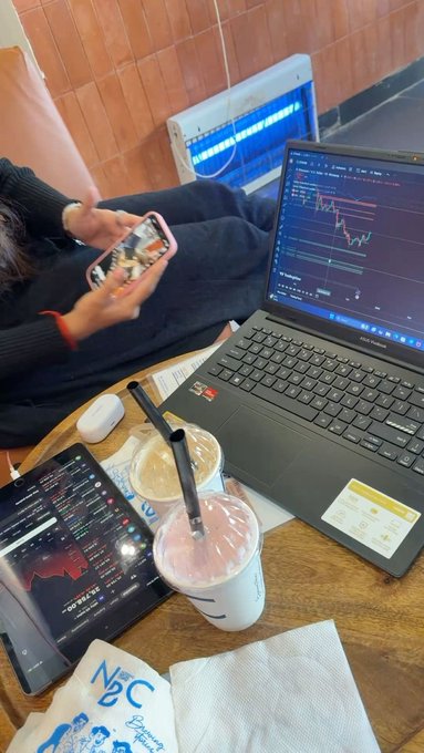 An overhead view of a coffee shop table showing two drinks (one coffee, one pink beverage), a tablet displaying stock charts, an open ASUS laptop also showing trading charts, a smartphone, and a person holding another phone in the background.