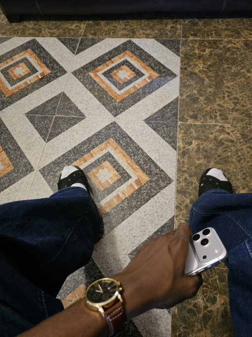 First-person view looking down at legs in jeans and white sneakers on a patterned tile floor, with one hand wearing a brown-strapped gold watch and holding a silver iPhone.