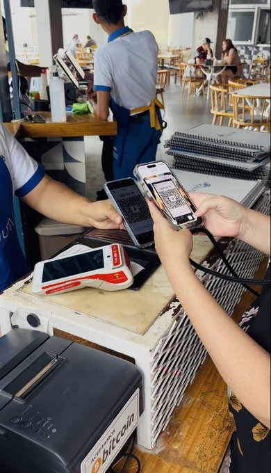 A customer scans a QR code on a phone at a restaurant counter to pay with Bitcoin, linking the post's "Ownership feels different @AquaBitcoin" theme to the real-world use of crypto for everyday transactions. The payment terminal and visible mobile screen highlight the seamless experience of using Bitcoin for in-person purchases.