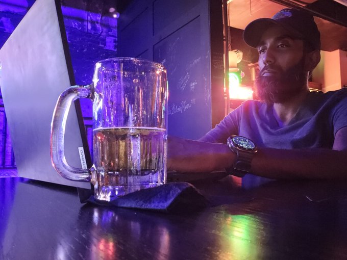 A bearded man wearing a dark baseball cap and t-shirt sitting at a wooden bar table with a large nearly-full glass mug of beer and a laptop in front of him, with purple and blue lighting in the background.