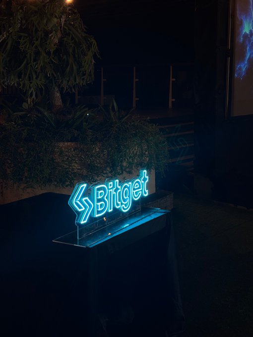A glowing blue neon 'Bitget' logo sign displayed on a stand at night, surrounded by plants.