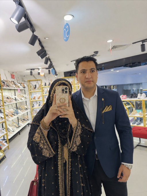 A couple takes a mirror selfie in a shoe store, with the woman in a black and gold embroidered hijab/abaya holding the phone and the man in a navy blazer standing beside her, sharing a stylish "GM" (Good Morning) greeting with their followers. A blue "02" sign is visible on the ceiling above them.