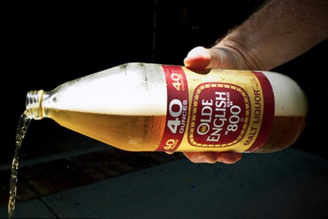 The image features a person pouring beer from an Old English 800 bottle, which has been opened and is now being poured out onto the ground. The scene takes place against a black background, making the beer bottle stand out prominently. The text "RIP to the homies who FOMO'd into XLM" appears in the image as well.