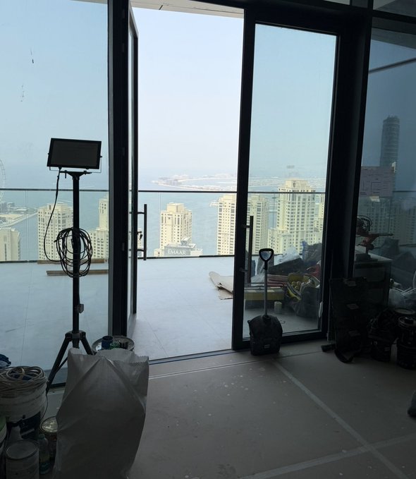 The image shows an unfinished penthouse interior with construction equipment, work lights on tripods, and scattered supplies, overlooking the Dubai Marina skyline with the "EMAAR" logo visible on a nearby building. The view from the balcony—featuring iconic high-rises and the waterfront—connects directly to the post's narrative of financial success through Bitcoin trading, illustrating the luxurious Dubai penthouse lifestyle made possible by cryptocurrency gains.