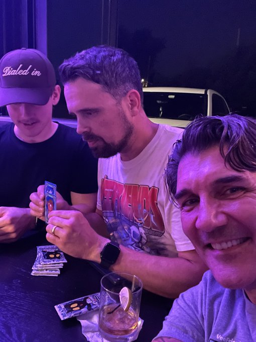 Three men at a nighttime gathering, with one wearing a 'Dialed in' cap and another in a Freaks t-shirt holding a card, while a third man smiles at the camera in the foreground.
