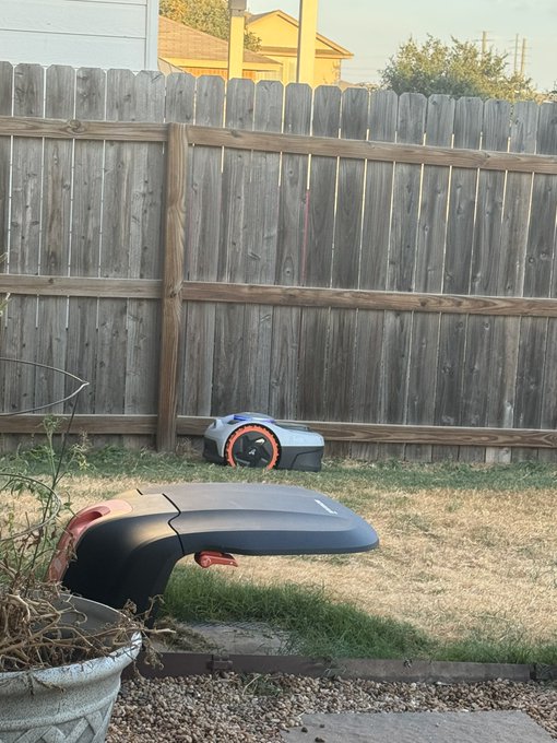 A robotic lawn mower sits in a backyard against a wooden fence, seemingly taking over the lawn-cutting duties while the owner kicks back to listen to music—nothing but space vibes for this automated chore.