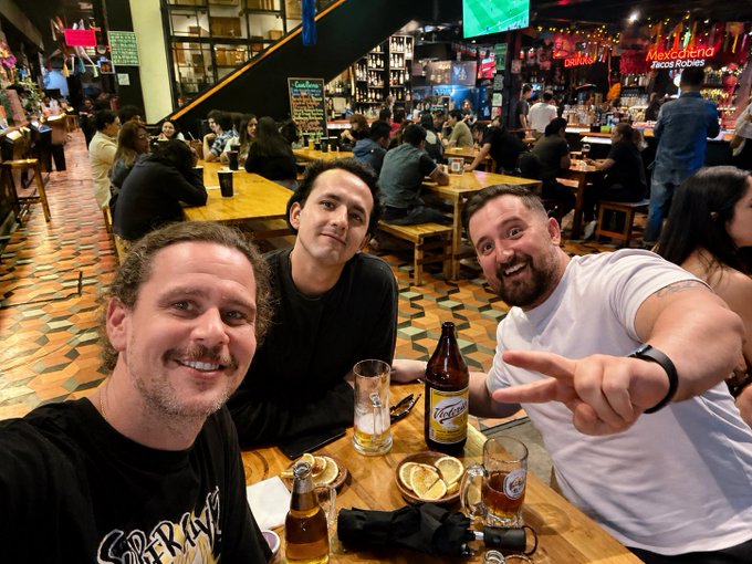 Three men take a selfie at a lively restaurant with wooden tables; the man on the right makes a peace sign, and beers and food are visible on their table.