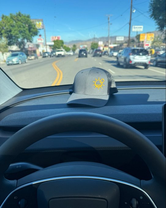 A gray baseball cap with a yellow bell pepper logo sits on a car's dashboard as the driver travels down a sunny street.