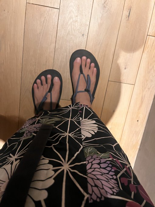 A first-person view looking down at feet wearing blue flip-flop sandals and a black floral patterned garment standing on a wooden floor.
