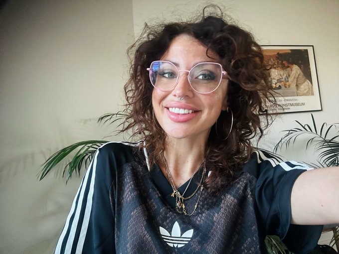 A smiling woman with curly hair and glasses wearing an Adidas shirt and layered necklaces, posing in a room with a framed poster.