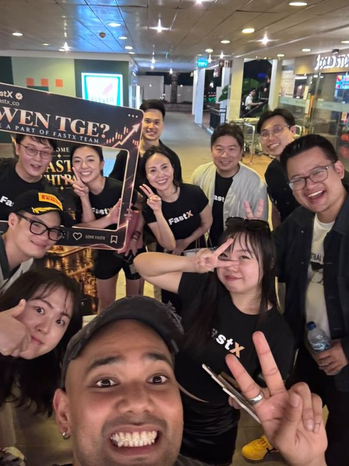 A group selfie of people wearing black 'FastX' t-shirts posing at an indoor venue, with one person holding a phone displaying a welcome frame reading 'WELCOME FASTX TEAM' and 'FASTX'.