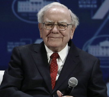 An elderly man with white hair wearing glasses, a dark suit, and red tie smiles while speaking at a microphone, appearing to be at a forum or conference panel.