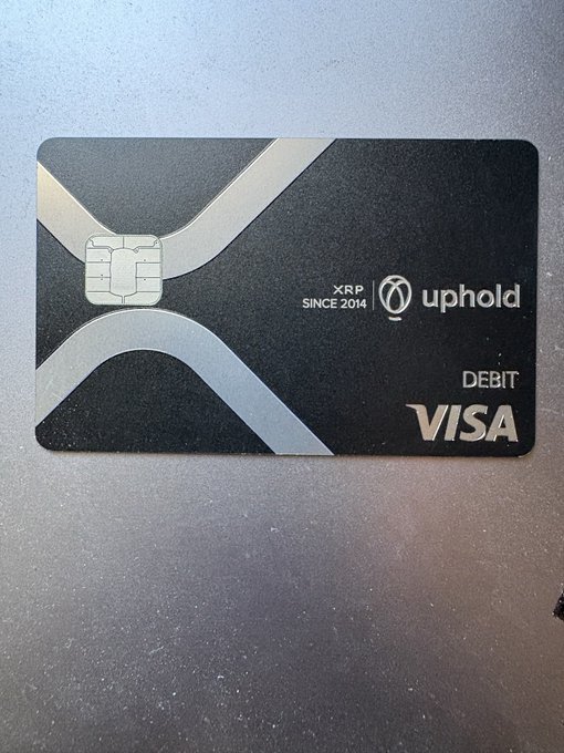 A black XRP debit Visa card from Uphold, marked "XRP SINCE 2014," featuring a silver chip and a metallic X design, resting on a dark surface.