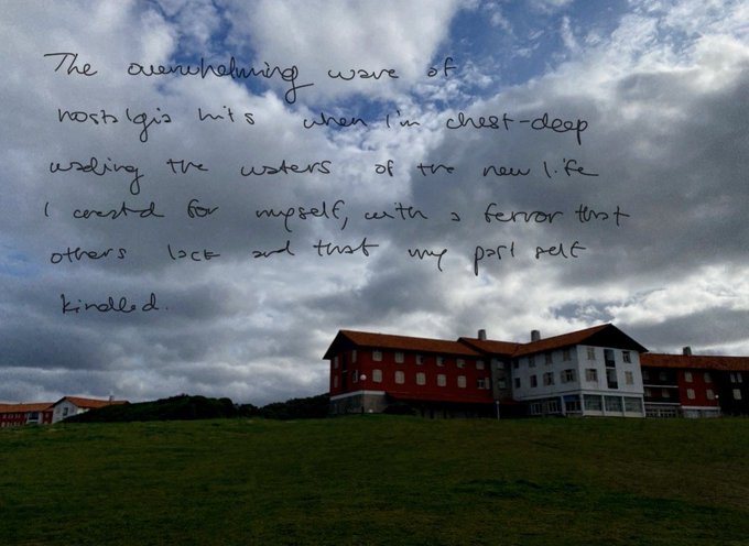 A landscape photo of red buildings with white trim on a green grassy hill under a cloudy sky, featuring handwritten text in the upper portion reading: "The overwhelming wave of nostalgia hits when I'm chest-deep wading the waters of the new life I created for myself, with a fervor that others lack and that my past self kindled."
