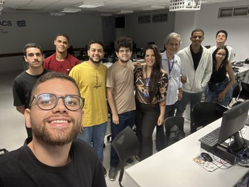 The image shows a group of people posing for a photo at their workplace or university. There are nine individuals in total, with one person wearing glasses and another person holding a computer mouse. The group appears to be gathered around a desk, which has a keyboard on top. They seem to be enjoying themselves as they pose together for the picture.