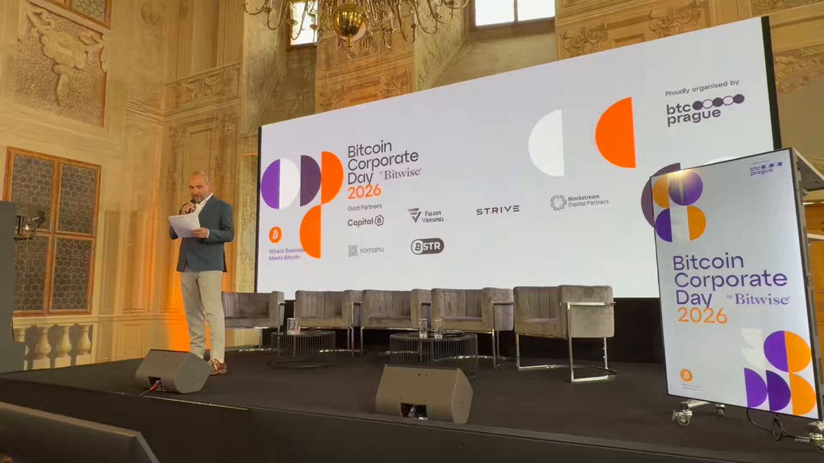 The image shows a man standing at a podium with a large screen behind him, giving a speech or presentation. The screen displays various logos and text related to Bitcoin Corporate Day 2016. There are several chairs placed around the stage, suggesting that this event may involve discussions or panel sessions. The man is dressed in a suit, indicating a formal setting for the event.