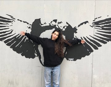A smiling woman wearing a black beanie and black sweatshirt stands in front of a mural with her arms spread, mimicking the outstretched wings of a bird painted on the wall behind her.