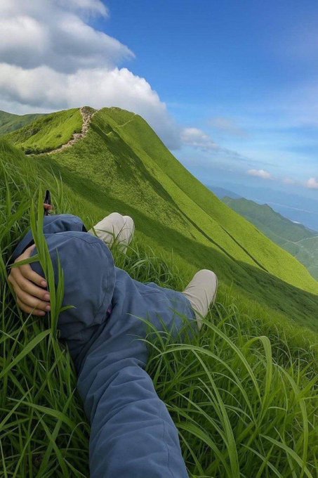Photograph taken from a low angle showing a person in a blue jacket lying on a grassy hillside with a winding dirt trail leading up a green mountain ridge under a partly cloudy sky.