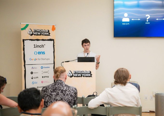 The image shows a man standing at a podium, giving a presentation or speech to an audience seated behind him. The room has a TV screen mounted on the wall above the podium, which displays a question for the attendees. There are several people sitting in chairs around the room, some of them wearing ties. A banner is also visible in the background, possibly displaying logos and information related to the event or organization.