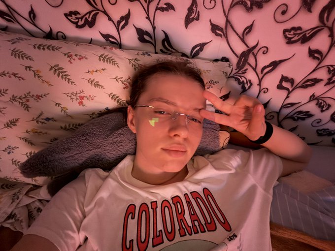 A selfie of a person wearing glasses, a white "COLORADO" t-shirt, and a heart filter, posing with a peace sign while lying on floral-patterned bedding.