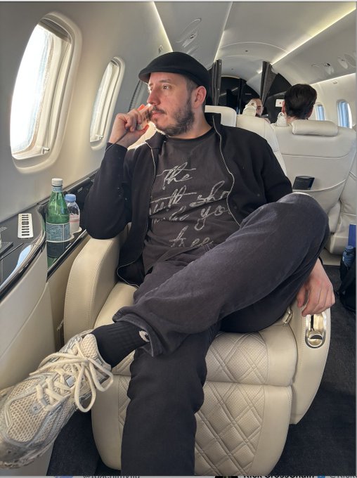 A man with a beard and mustache, wearing a black cap and jacket, sits in a white leather airplane seat, resting his hand near his mouth while looking out the window.