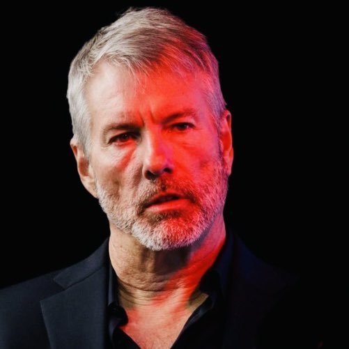 A middle-aged man with gray hair and a beard is shown in a close-up portrait against a dark background, illuminated by red lighting. The image accompanies breaking news about Michael Saylor's company Strategy selling an additional $102.4M in Bitcoin, bringing their 2026 total sales to $320.86 million.